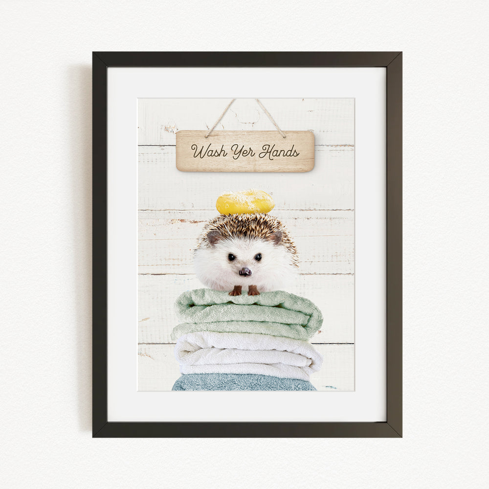 A framed image of a hedgehog sitting on a stack of towels, with a wooden sign above that says "Wash Your Hands".