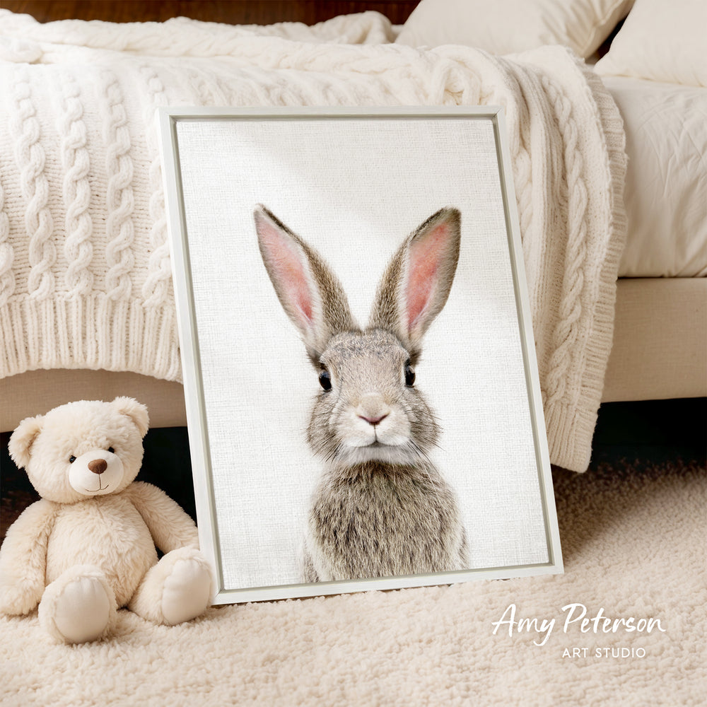 A framed image of a rabbit with large ears is displayed on a white bedspread, accompanied by a teddy bear on the floor.