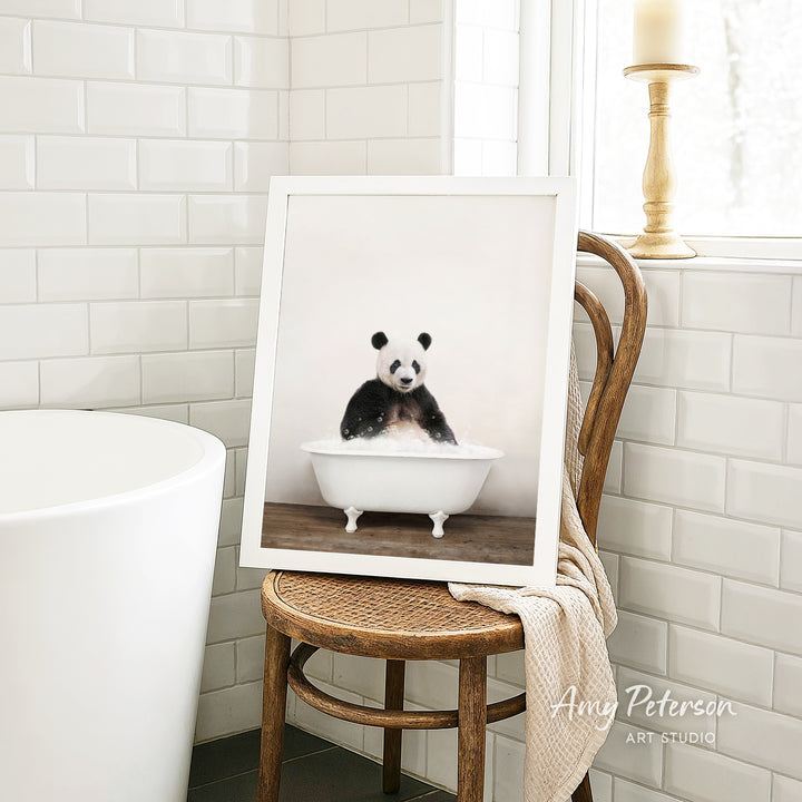 A framed image of a panda sitting in a white bathtub is displayed on a wooden chair in a bathroom setting.