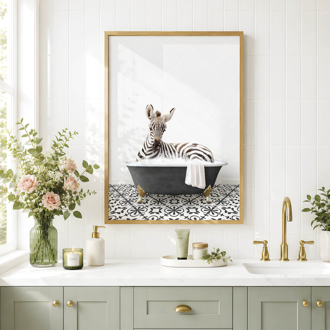 A framed image of a zebra sitting in a bathtub is displayed on a white tiled wall above a bathroom vanity with a sink, green cabinets, and a vase of flowers.