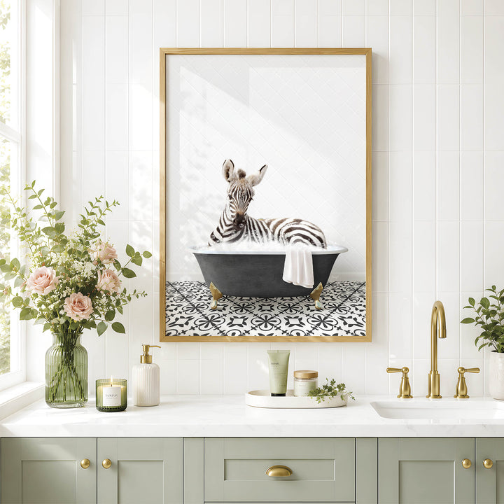 A framed image of a zebra sitting in a bathtub is displayed on a white tiled wall above a bathroom vanity with a sink, green cabinets, and a vase of flowers.