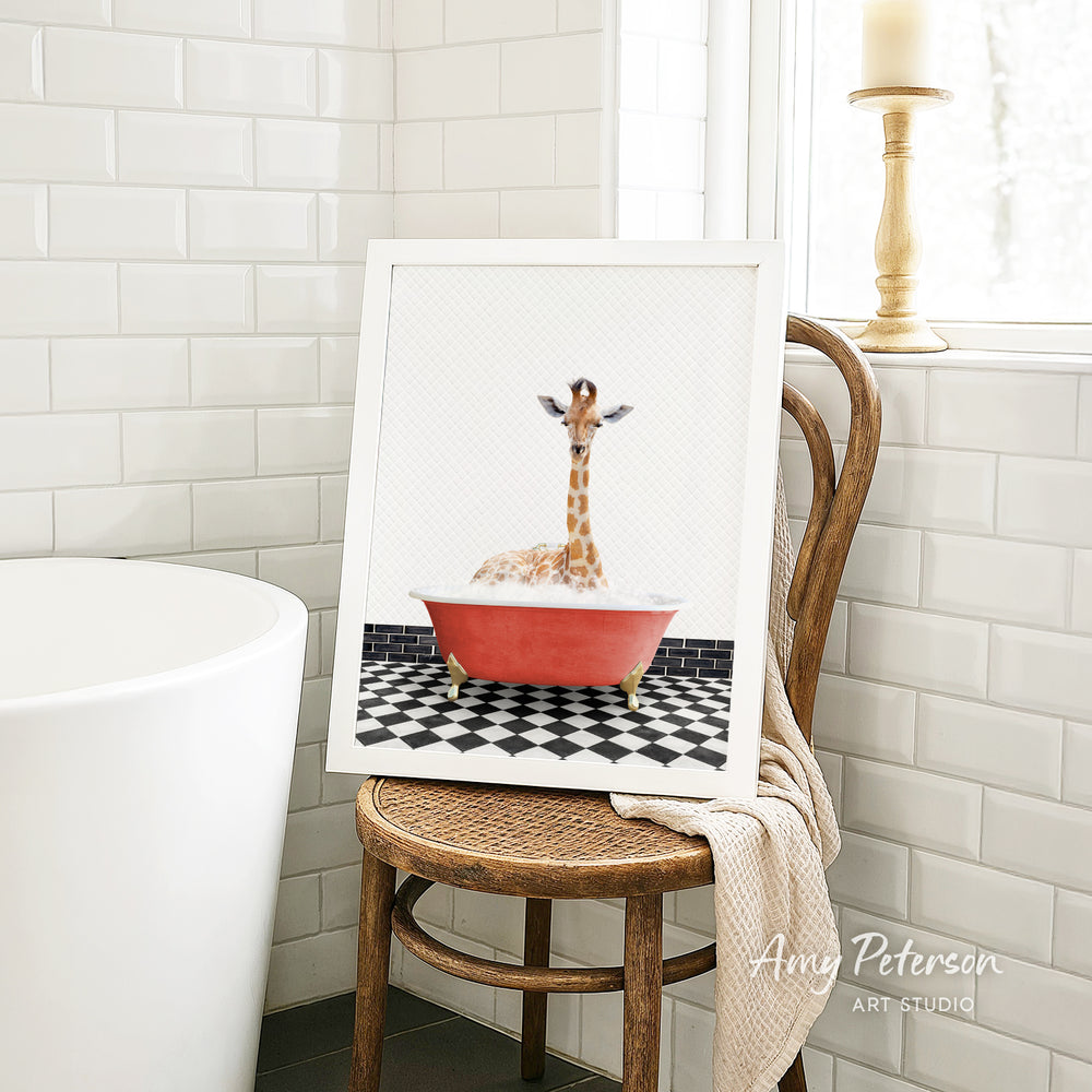 A framed image of a giraffe sitting in a red bathtub, surrounded by a black and white checkered floor, is displayed on a wooden chair in a bathroom setting.