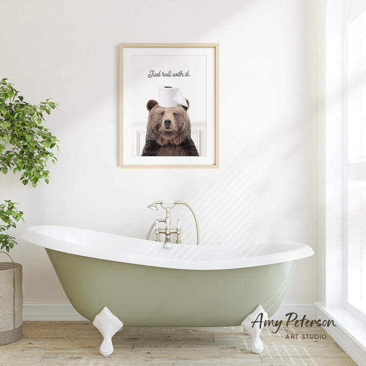 A framed image of a brown bear wearing a toilet paper hat is displayed on a white wall above a green clawfoot bathtub in a bright, minimalist bathroom.