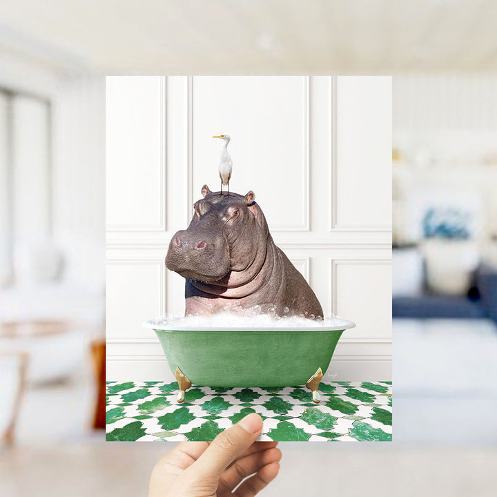 A hand holds up a framed image of a hippopotamus sitting in a green bathtub, with a seagull perched on its head.