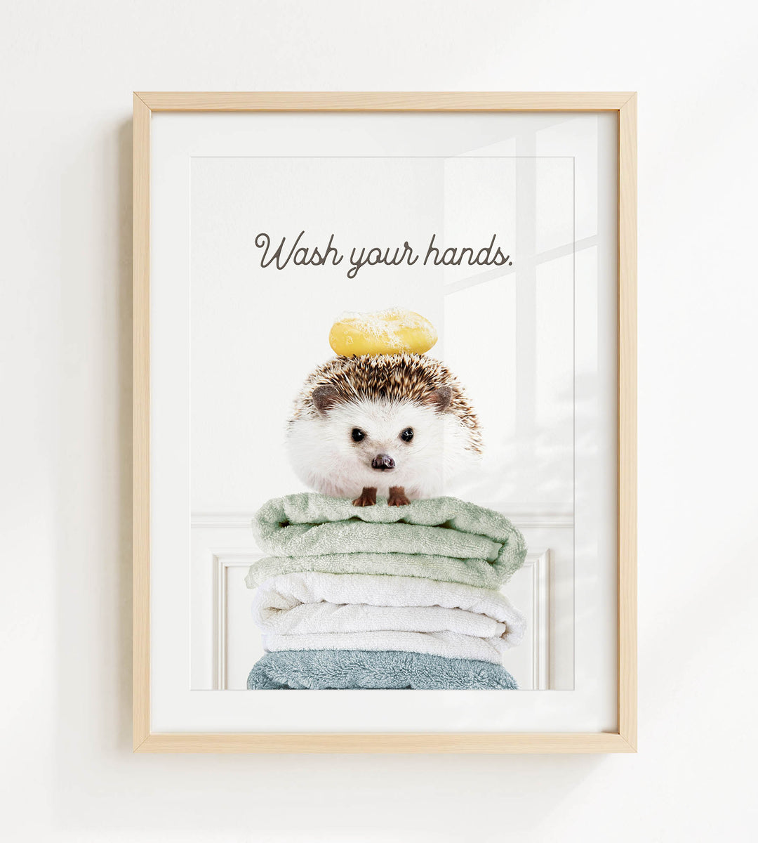 A framed image of a hedgehog sitting atop a stack of towels, with the text "Wash your hands" written above it.