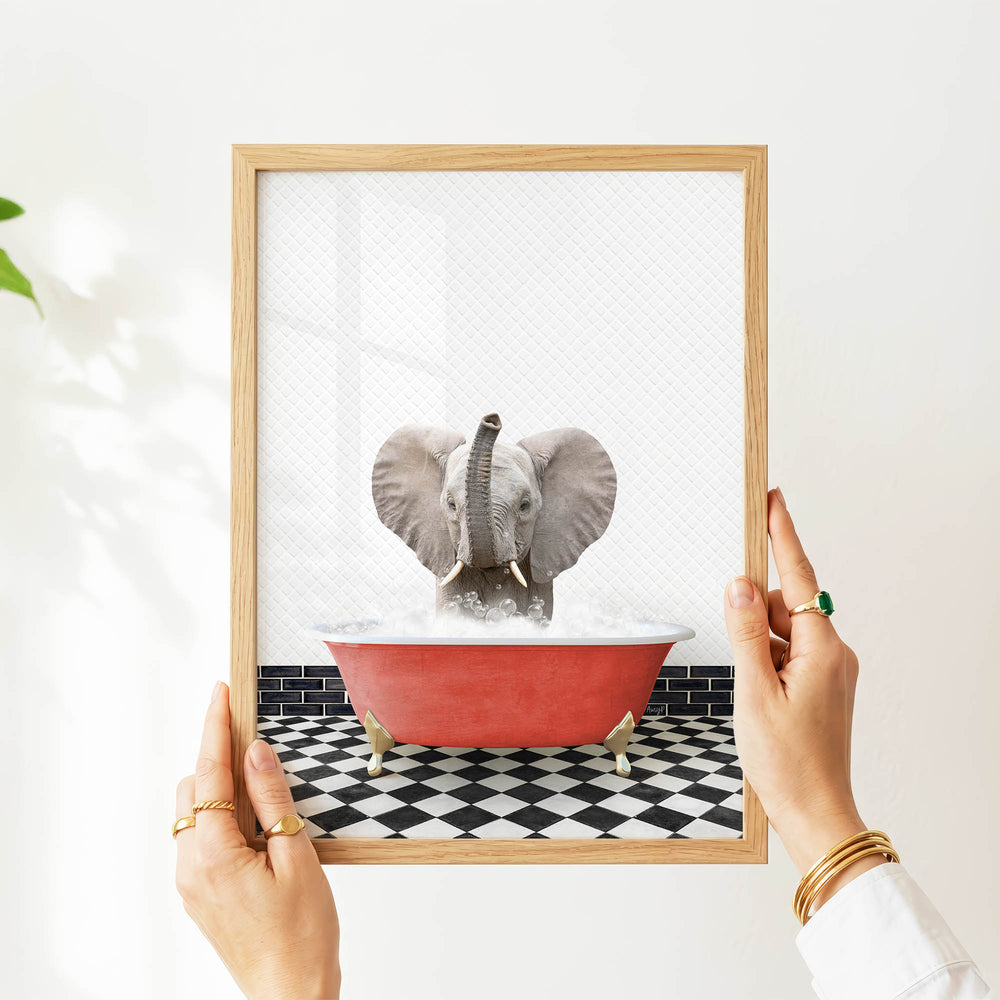 A person's hands are holding a framed picture of a gray elephant sitting in a red bathtub filled with water, surrounded by a black and white checkered floor.