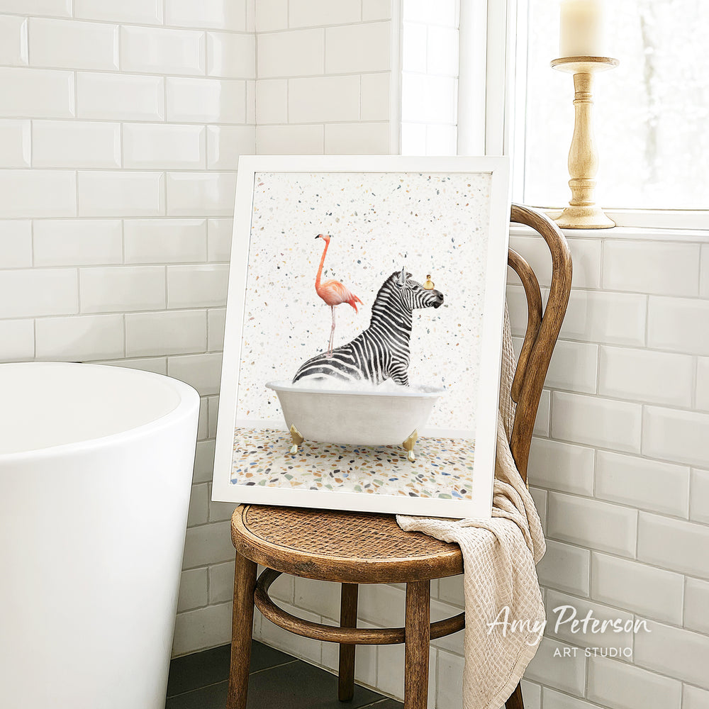 A framed image of a zebra sitting in a bathtub, with a pink flamingo standing on its back, is displayed on a wooden chair in a bathroom setting.