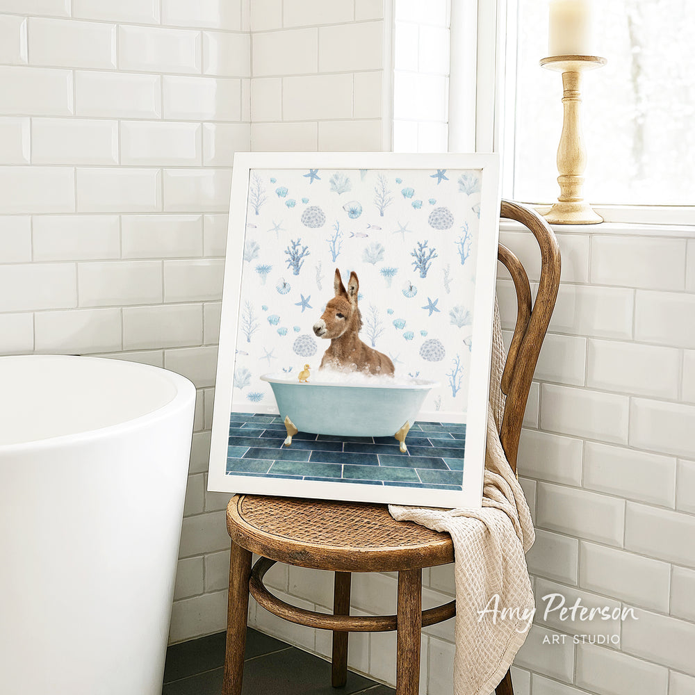 A framed illustration of a dog sitting in a bathtub, surrounded by blue and white floral patterns, is displayed on a wooden chair in a bathroom setting.