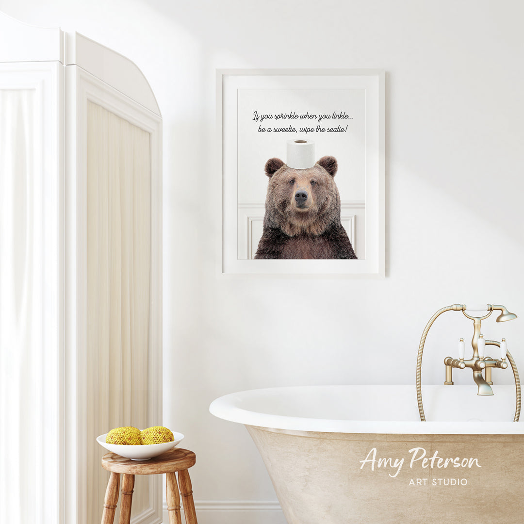 a bathroom with a framed picture of a bear sitting on a roll of toilet paper, and a wooden stool with a bowl of fruit in front of it.