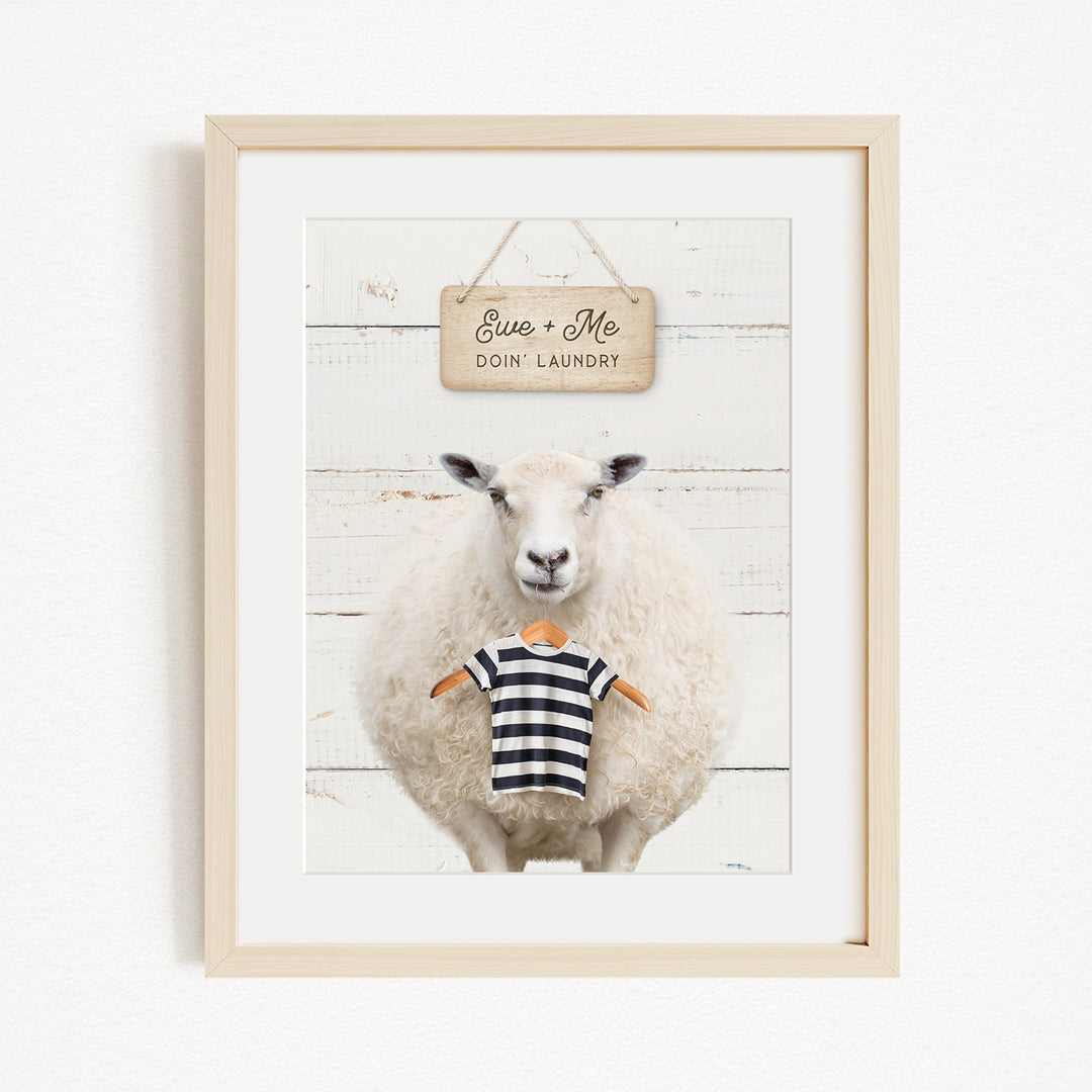A framed image of a sheep wearing a striped shirt, with a sign above it that says "Ewes + Me Doin Laundry".