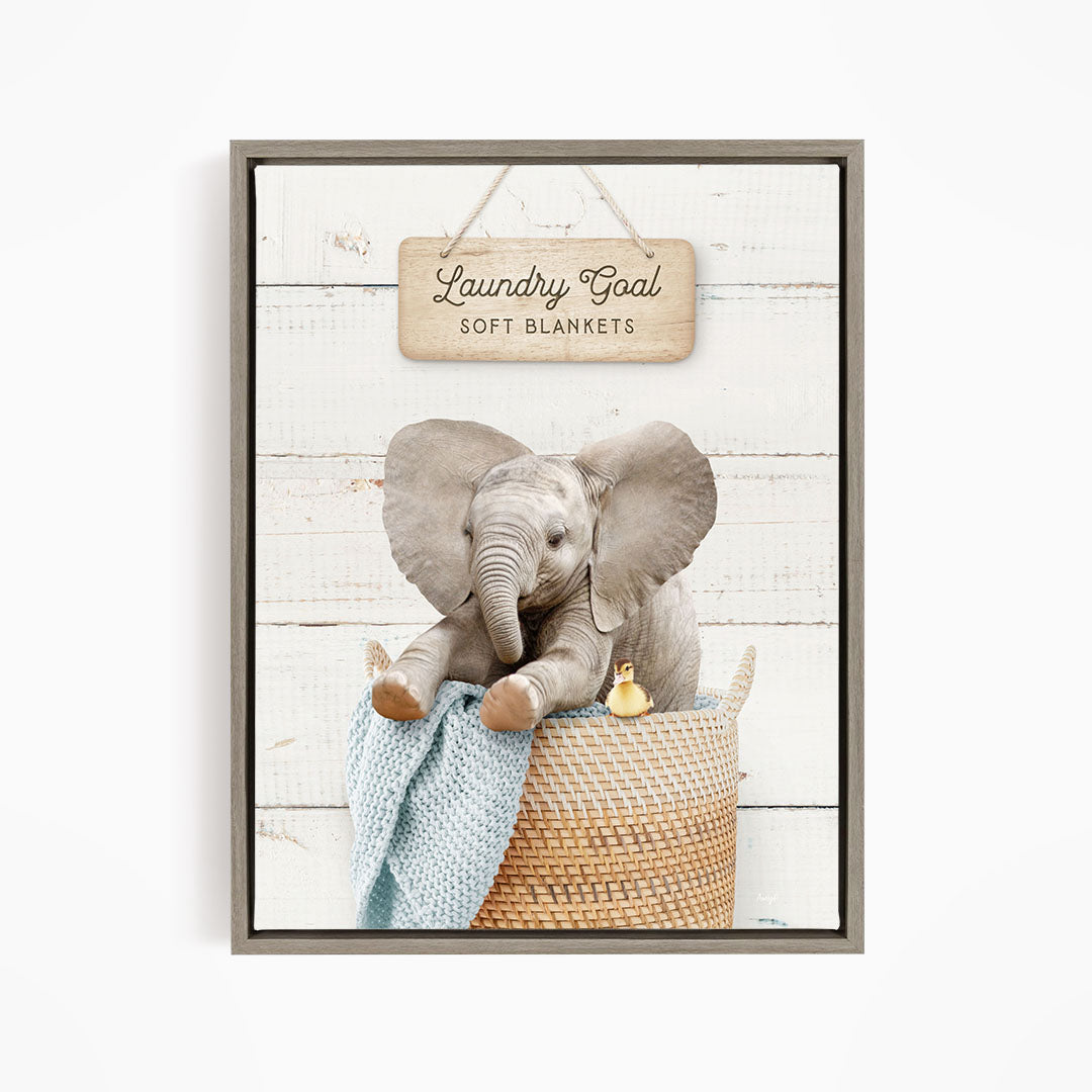 A framed image of an elephant sitting in a basket, with a sign above it that says "Laundry Goal Soft Blankets".