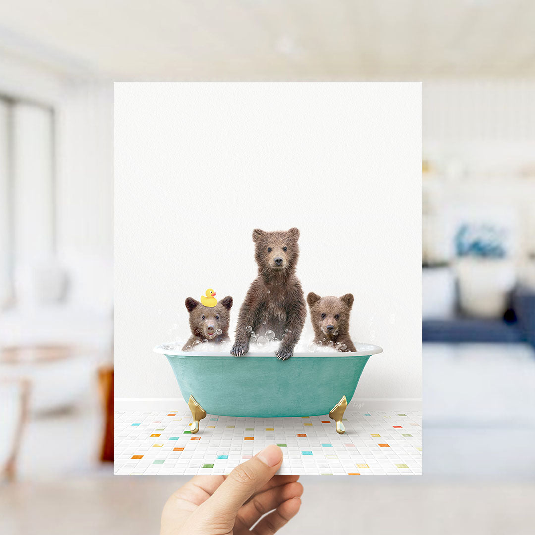 A hand holds up a framed image depicting three adorable brown bears sitting in a green bathtub filled with water, surrounded by colorful tiles on the floor.
