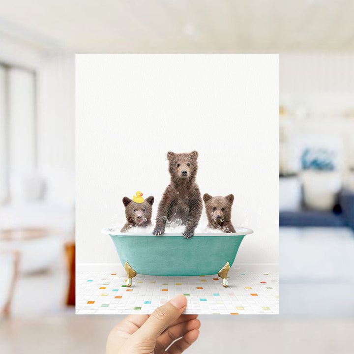 A hand holds up a framed image depicting three adorable brown bears sitting in a green bathtub filled with water, surrounded by colorful tiles on the floor.