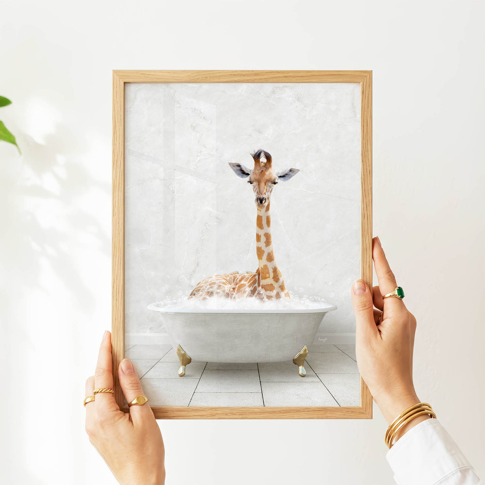 A person's hands are holding a framed picture of a giraffe sitting in a bathtub filled with water.