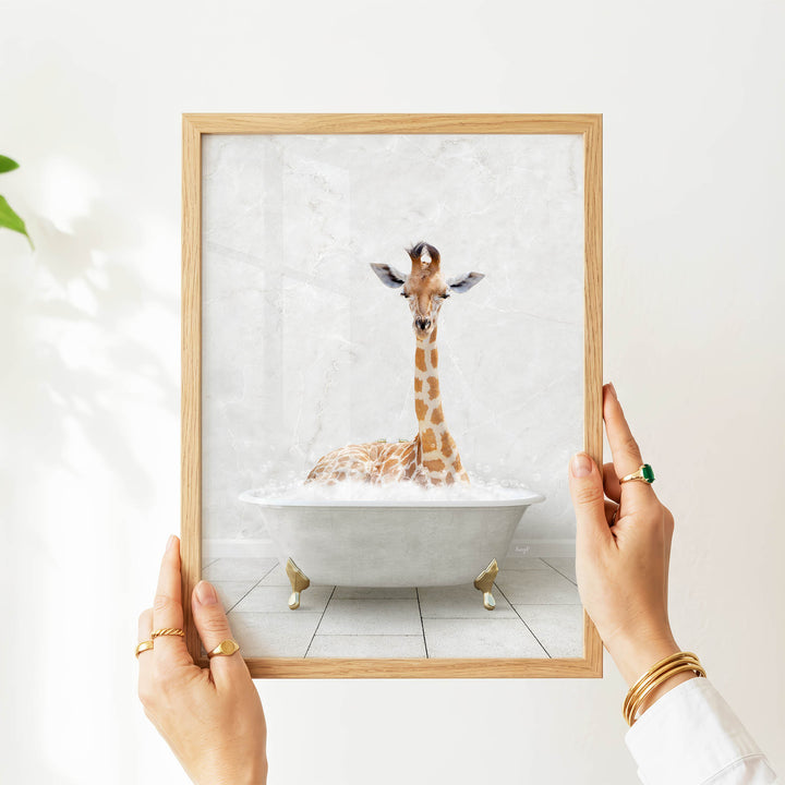 A person's hands are holding a framed picture of a giraffe sitting in a bathtub filled with water.