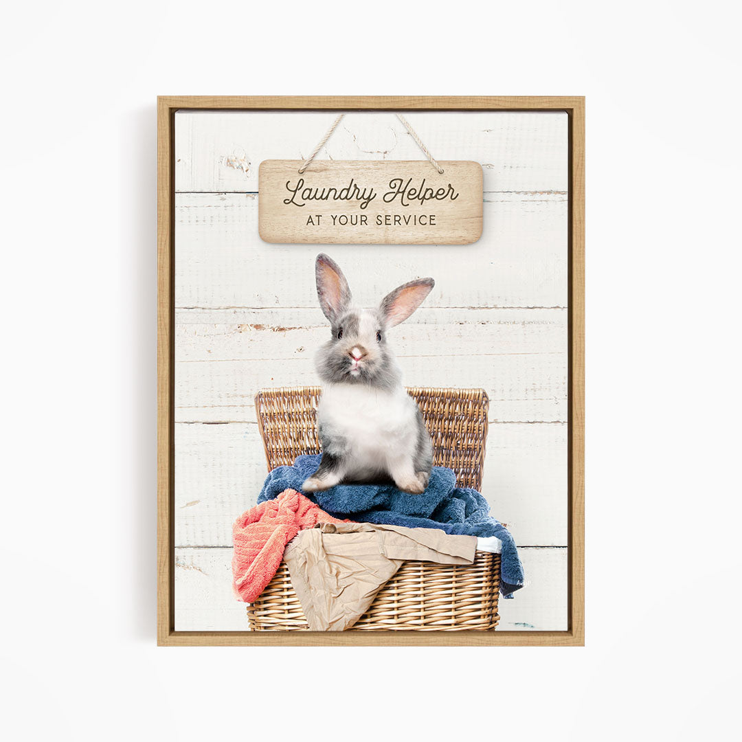A gray and white rabbit sitting in a wicker laundry basket, with a wooden sign above that reads "Laundry Helper at your service".