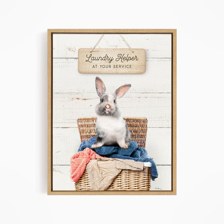 A gray and white rabbit sitting in a wicker laundry basket, with a wooden sign above that reads "Laundry Helper at your service".