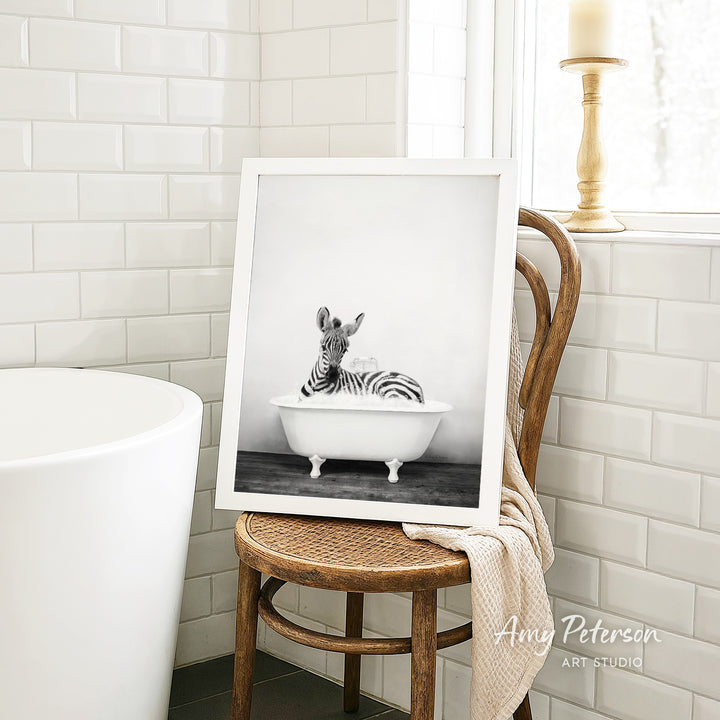 A framed black and white photograph of a zebra sitting in a bathtub, with a towel draped over the back of a wooden chair in a bathroom setting.