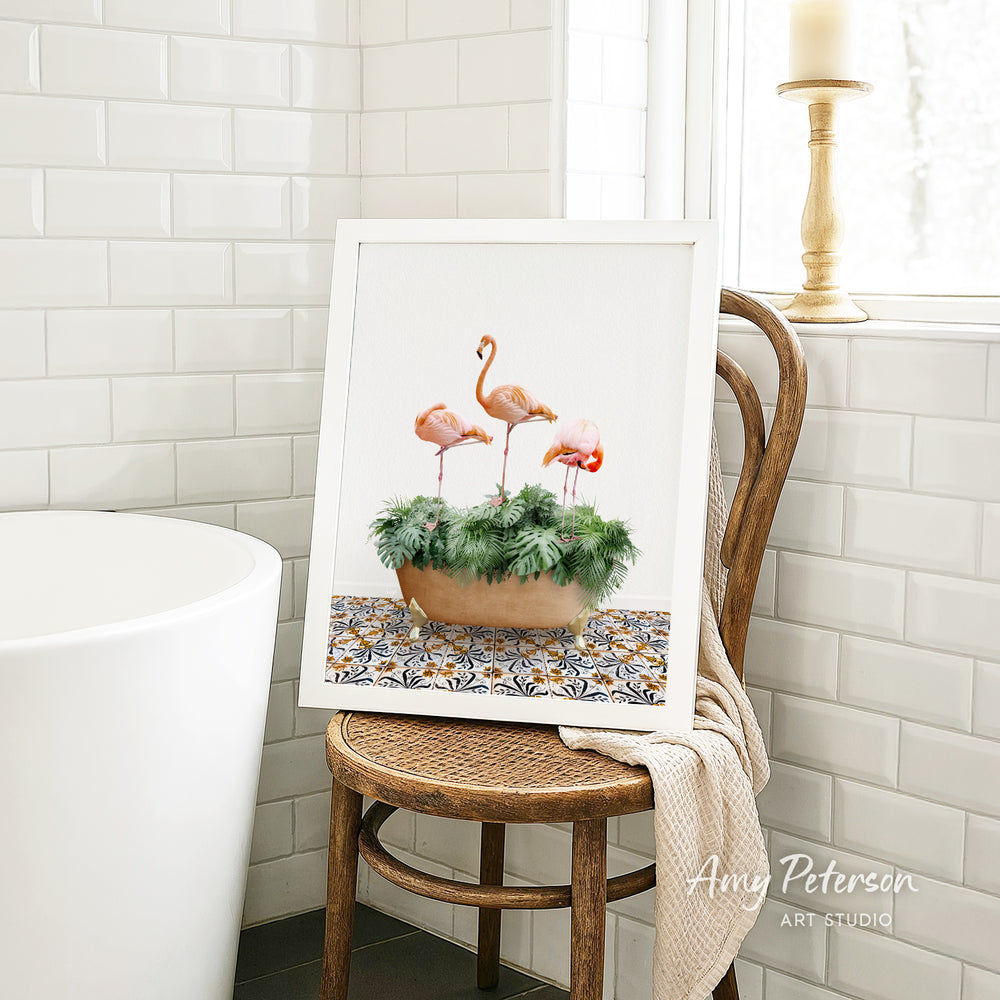 A framed painting of three flamingos standing on a green plant is displayed on a wooden chair in a bathroom setting.