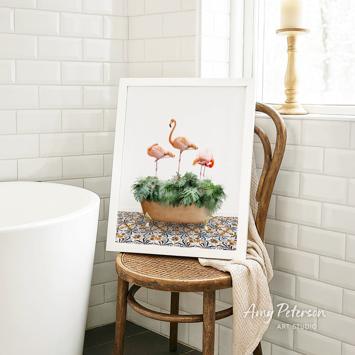 A framed painting of three flamingos standing on a green plant is displayed on a wooden chair in a bathroom setting.