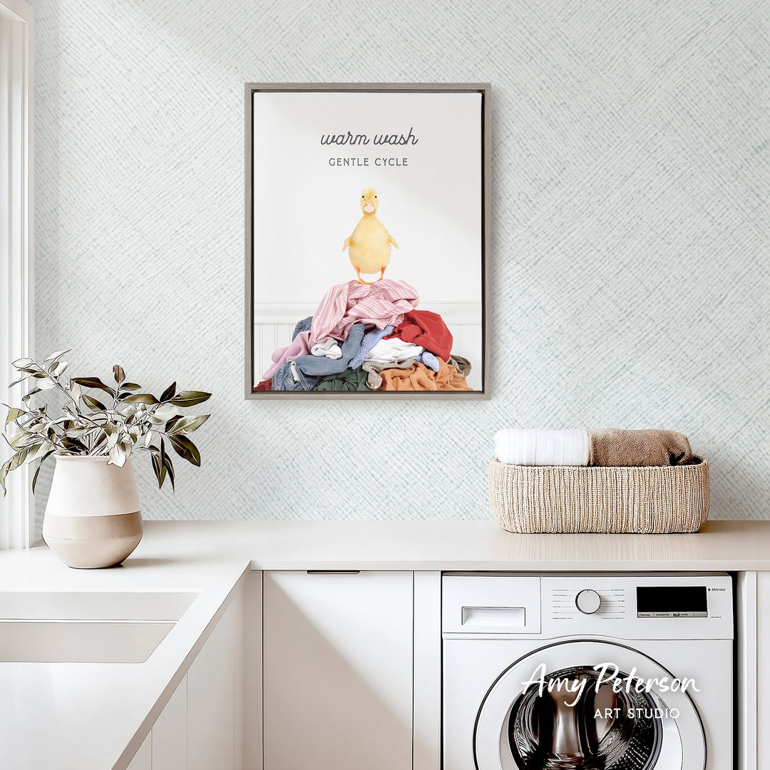 a framed poster of a yellow duck on a pile of laundry, hanging on a wall above a white countertop with a basket of folded clothes and a potted plant.