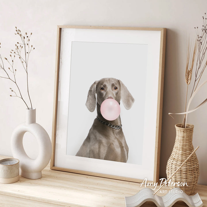 A framed photograph of a dog blowing a bubble with a pink bubble gum bubble, accompanied by a wicker vase and a white ceramic vase on a wooden surface.