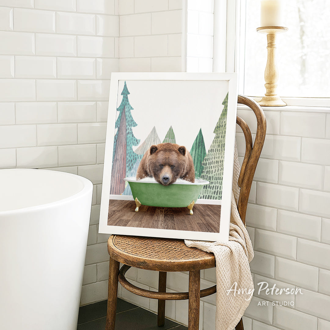 A framed image of a brown bear sitting in a green bathtub, surrounded by a forest scene, is displayed on a wooden chair in a bathroom setting.