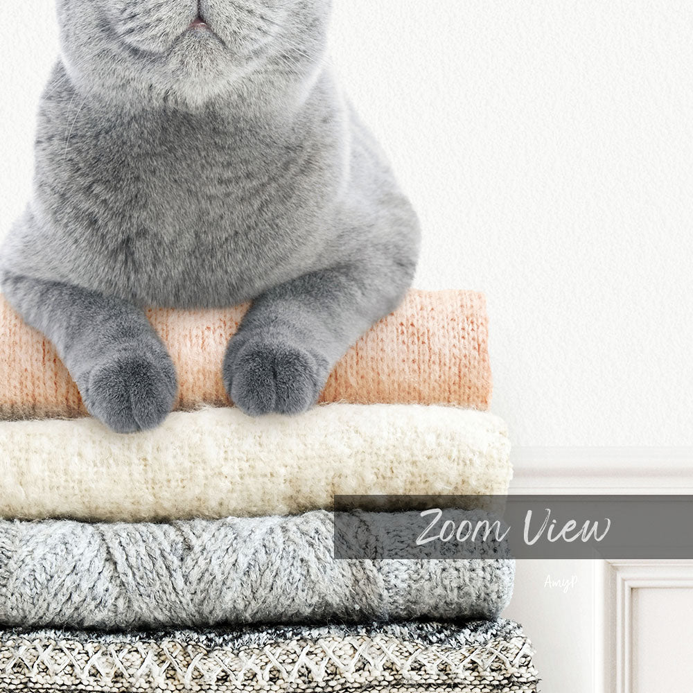 A gray cat sitting on top of a stack of variously colored sweaters.