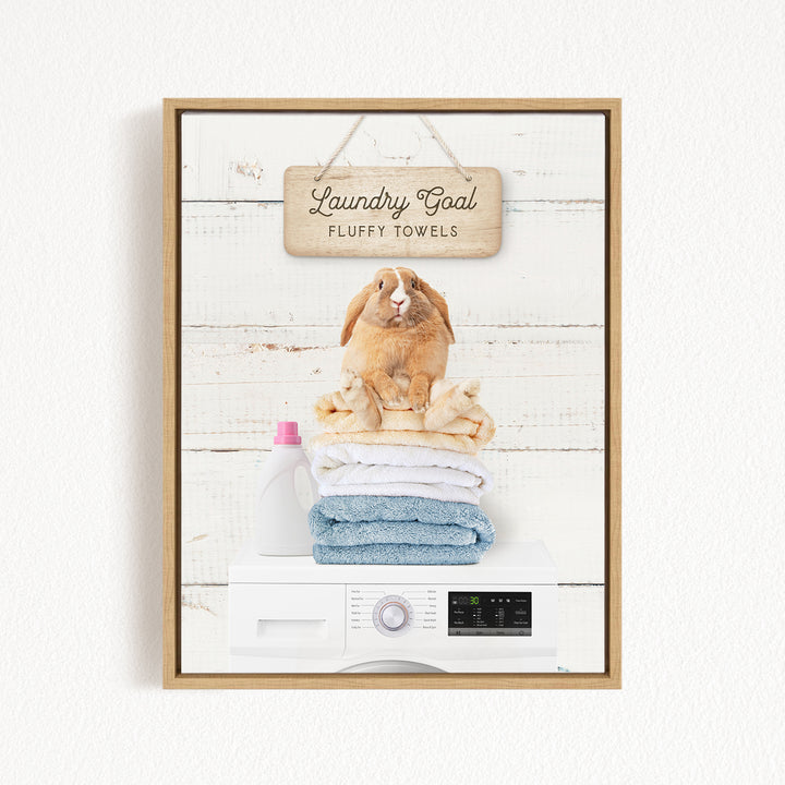 A wooden sign with the text "Laundry Goal Fluffy Towels" hangs above a stack of folded towels and a washing machine.