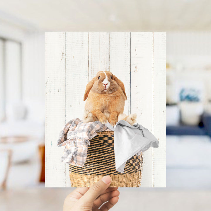 A person's hand is holding up a photograph of a brown rabbit sitting in a woven basket, which is placed on a white wooden surface.