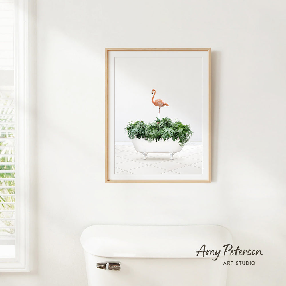 A framed print of a flamingo standing on a plant in a white bathtub is hanging on a white wall.
