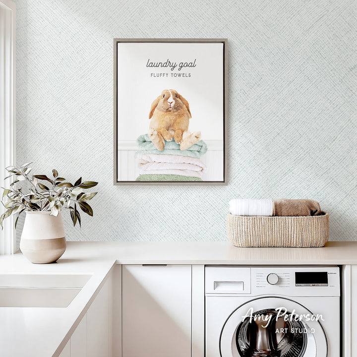 a laundry room with a framed poster of a rabbit sitting on a pile of fluffy towels, a basket of folded towels, and a potted plant on the counter.