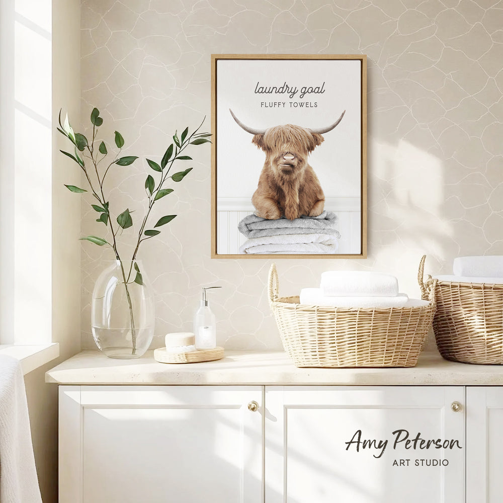 a framed poster of a Highland cow with horns, hanging on a wall above a white cabinet with baskets and a vase of leaves.