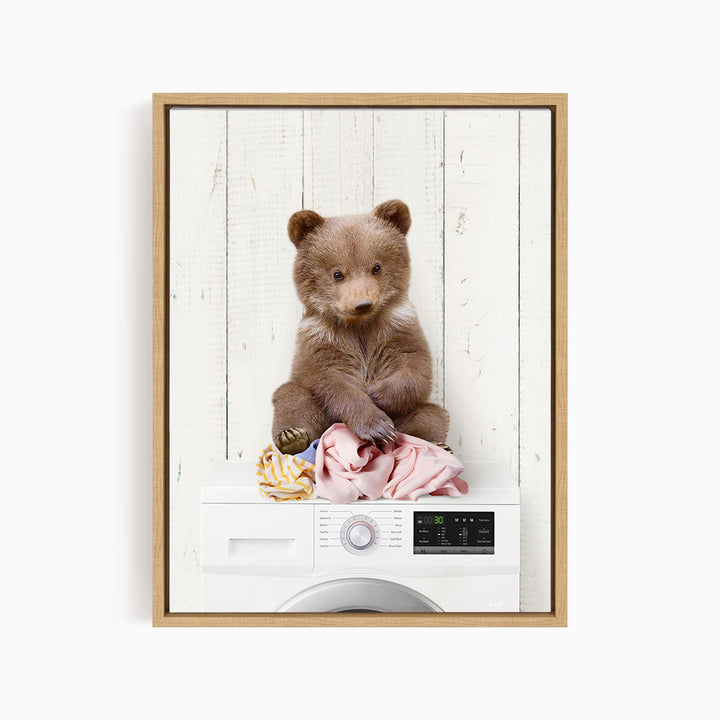 A brown bear sitting on top of a white washing machine, with clothes hanging on the machine.