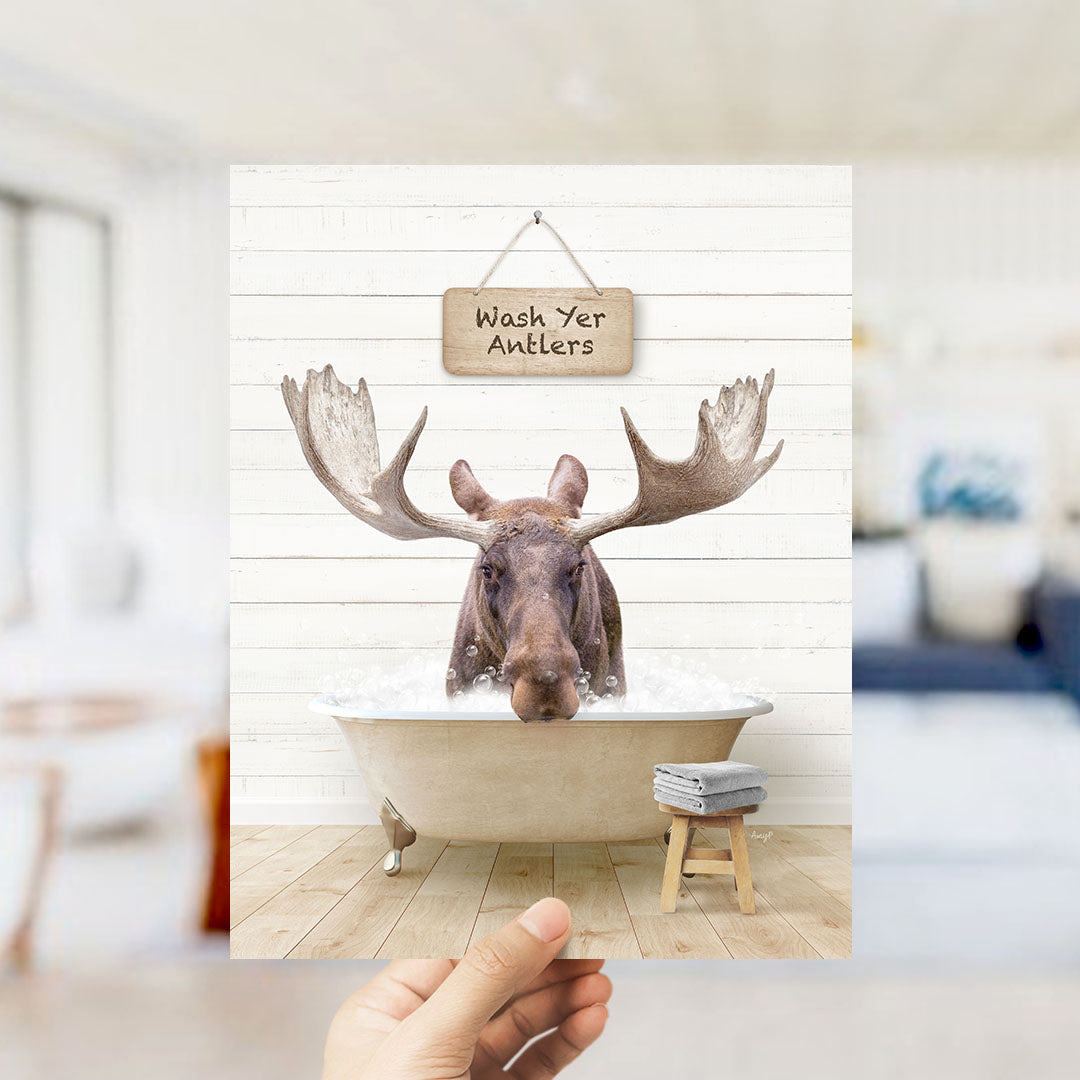 A moose with large antlers is sitting in a bathtub filled with water, with a sign above it that says "Wash Yer Antlers".