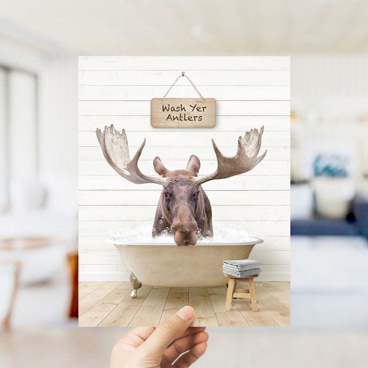 A moose with large antlers is sitting in a bathtub filled with water, with a sign above it that says "Wash Yer Antlers".