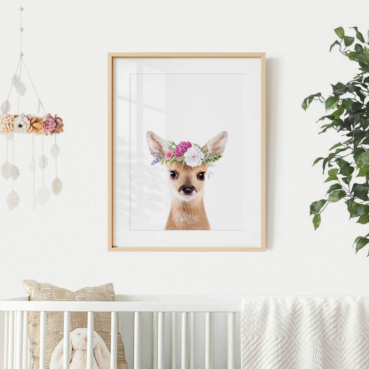 A framed picture of a deer wearing a flower crown is hanging on a white wall, with a crib and a hanging mobile with flowers in the background.