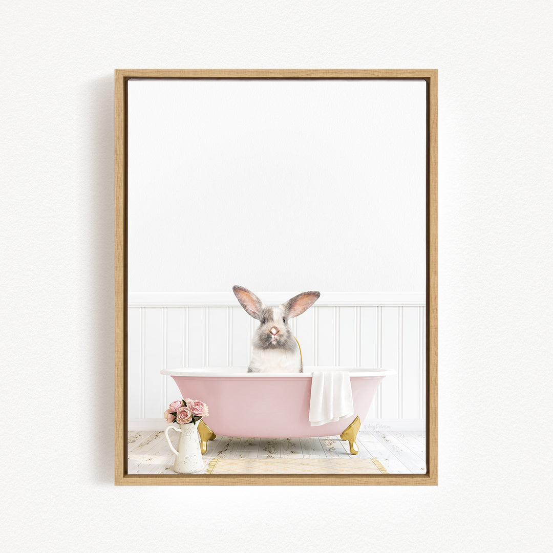 A gray rabbit sitting in a pink bathtub, surrounded by a white towel and a vase of flowers, in a white-walled bathroom.