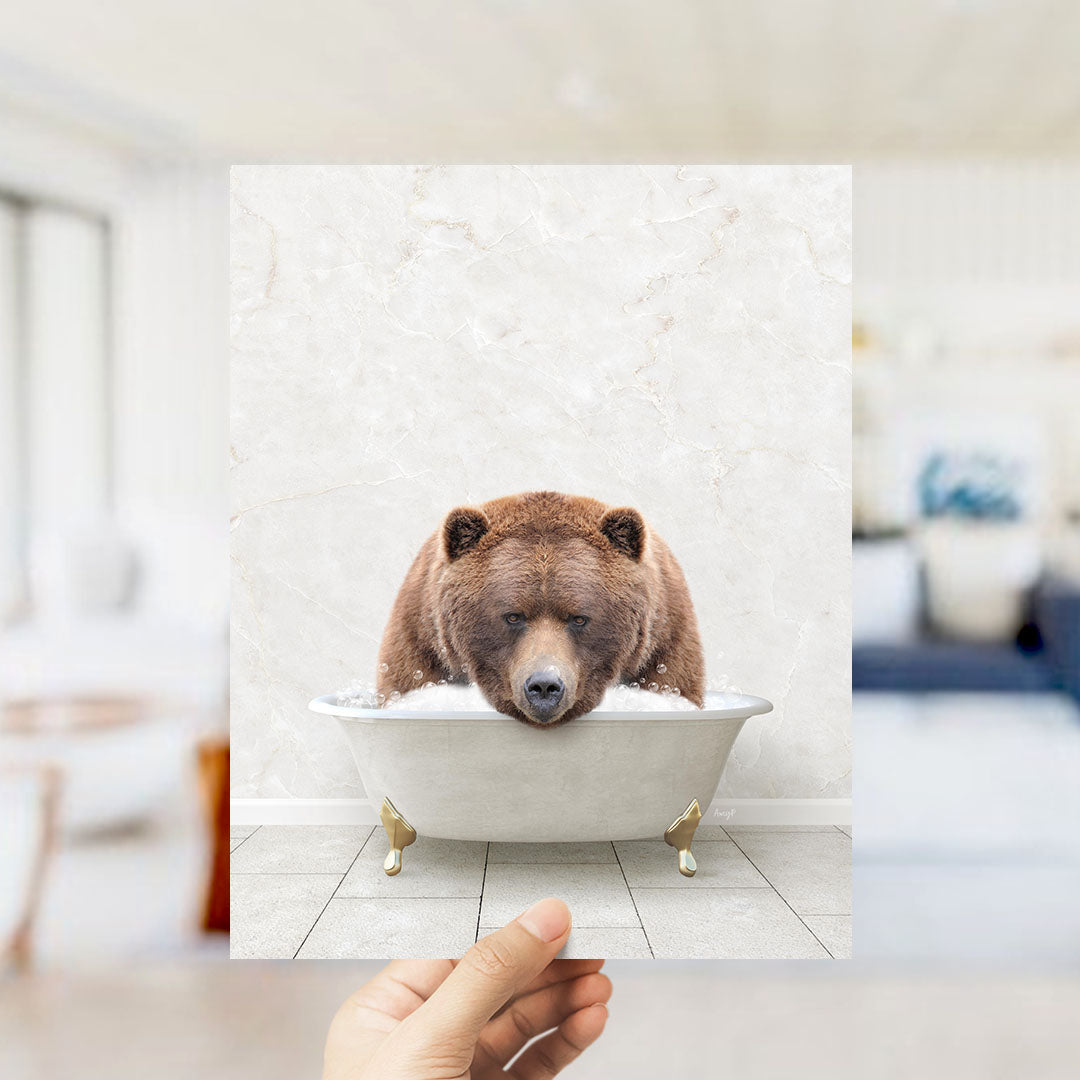 A hand holds up a framed image of a brown bear sitting in a bathtub filled with water, creating a whimsical and playful scene.
