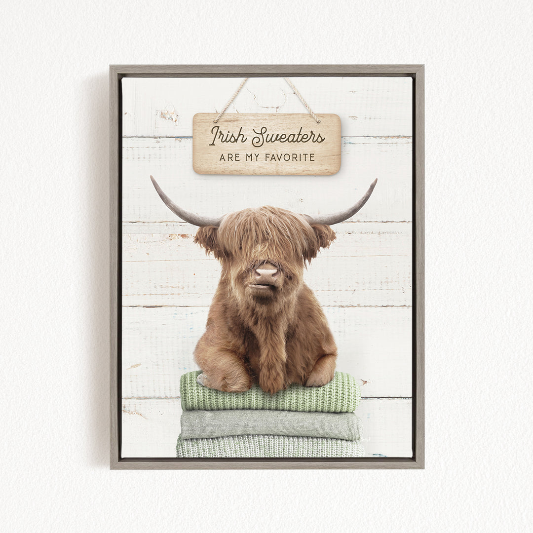 A brown cow with long horns is sitting on a stack of green blankets, with a sign above that says "Irish Sweaters are my favorite".