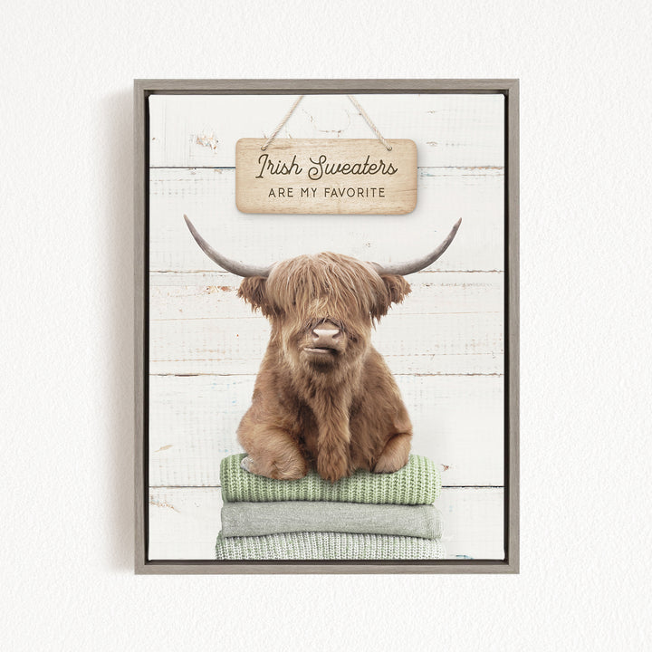 A brown cow with long horns is sitting on a stack of green blankets, with a sign above that says "Irish Sweaters are my favorite".