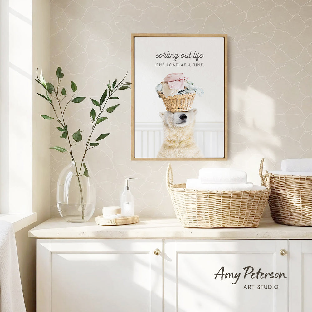 a framed poster of a polar bear with a basket on its head, hanging on a wall above a white cabinet. The cabinet has two woven baskets on top. There is also a vase with a plant and a soap dispenser on the cabinet. The text "sorting out life one load at a time" is written above the polar bear in the poster.