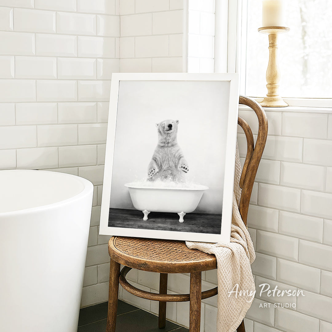 A framed black and white photograph of a polar bear sitting in a bathtub, with a towel draped over the back of a wooden chair in a bathroom setting.