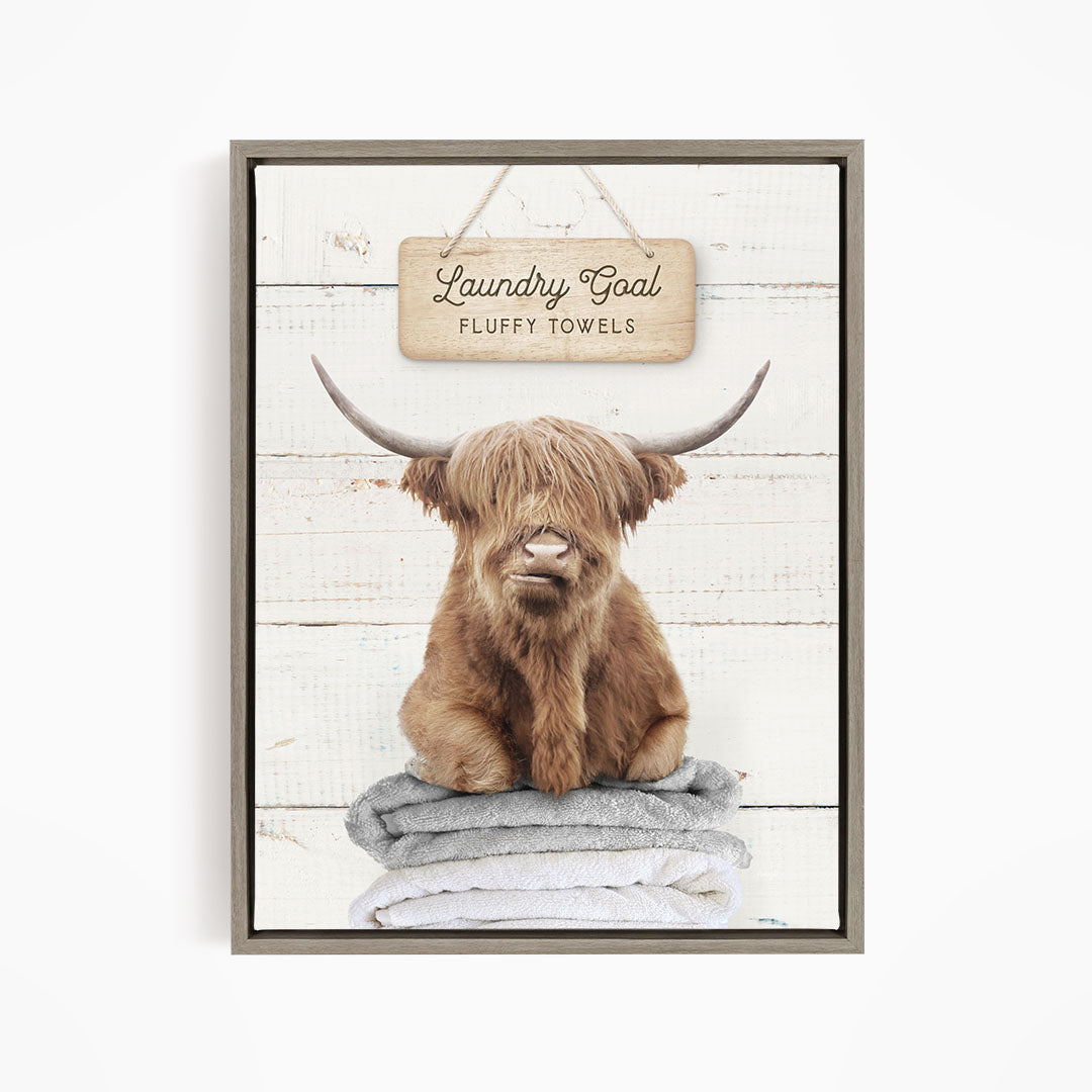A framed image of a brown cow with long horns sitting on a stack of fluffy towels, with a sign above that says "Laundry Goal Fluffy Towels".