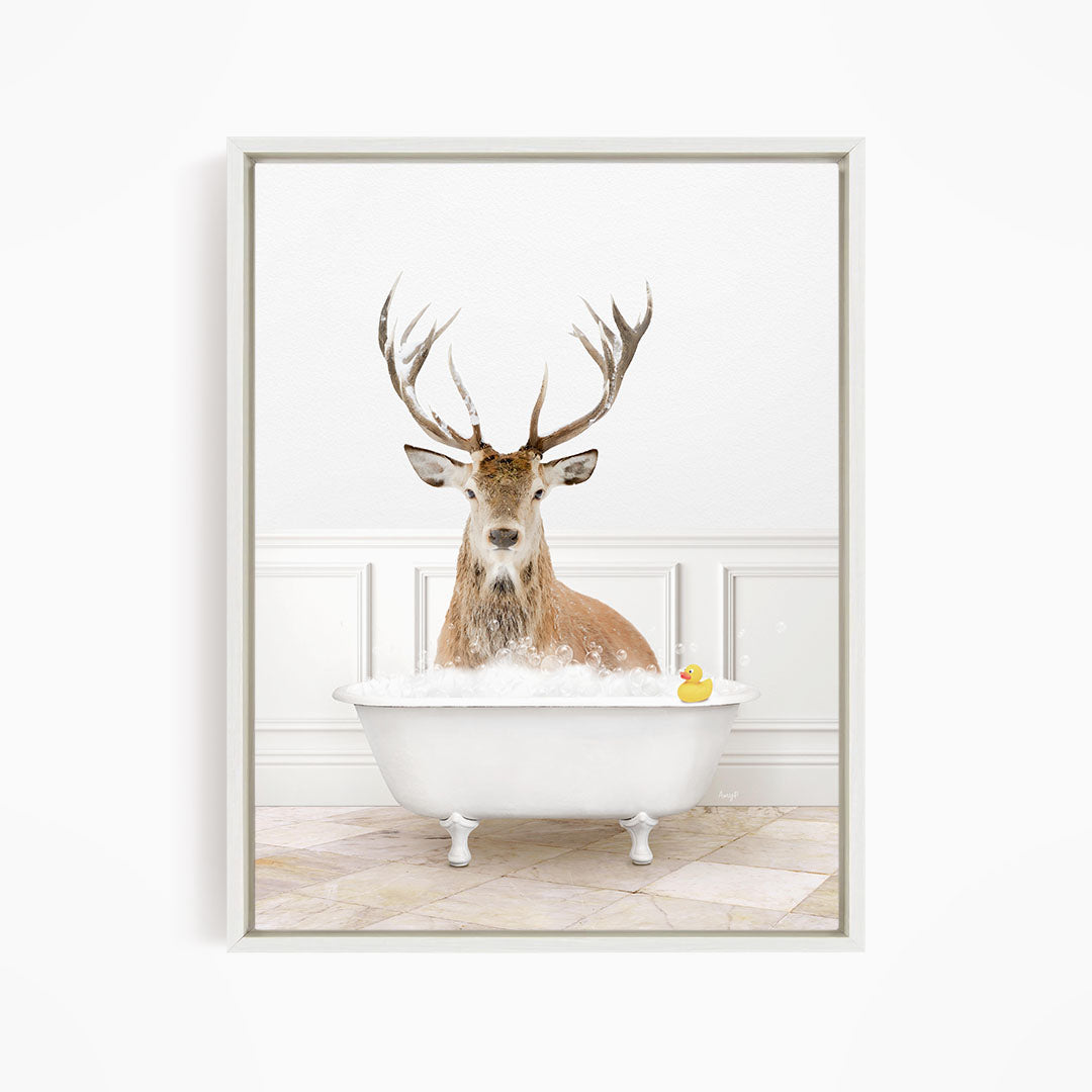 A deer with large antlers is sitting in a white bathtub filled with bubbles, with a yellow rubber duck floating in the water.