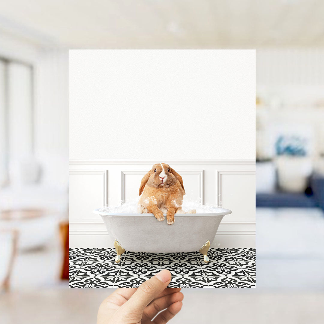A hand holds up a photograph of a brown rabbit sitting in a white bathtub filled with bubbles, surrounded by a black and white patterned floor.