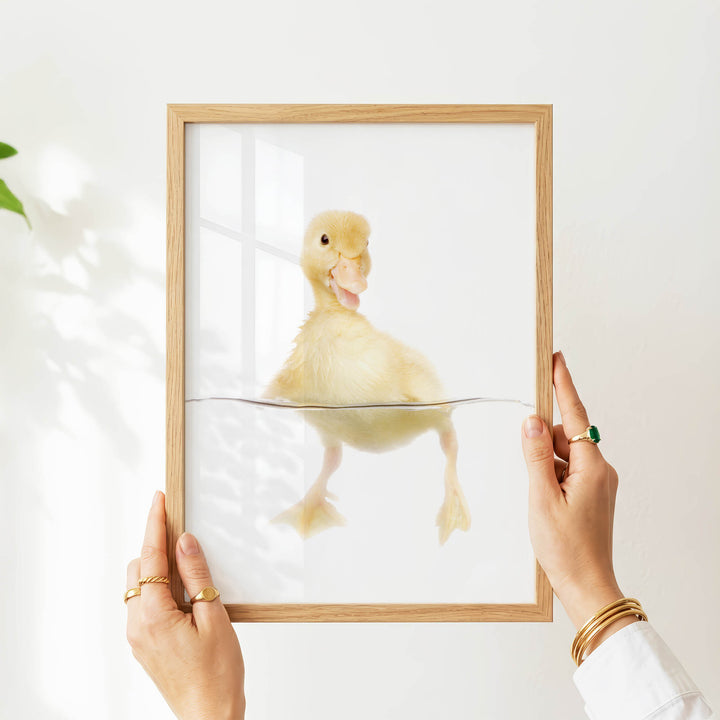 A person's hands are holding a picture frame with a photograph of a yellow duck inside it, against a white background.
