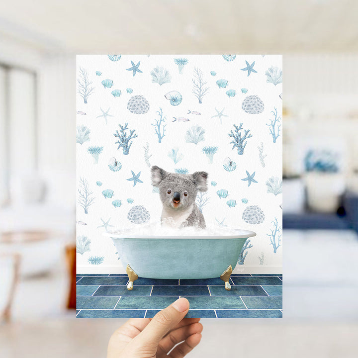 A hand is holding up a framed image of a koala bear sitting in a bathtub filled with bubbles, surrounded by a blue and white ocean-themed wallpaper.