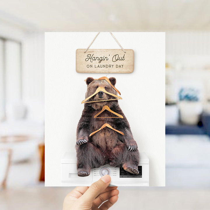 A hand holds up a framed image of a bear sitting on a washing machine, with a wooden sign above it that says "Hangin' Out on laundry day".