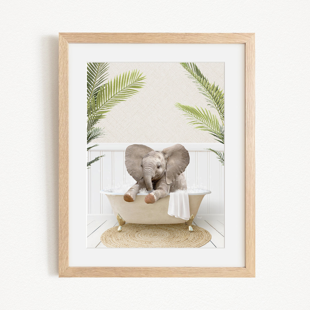 A framed image of a baby elephant sitting in a bathtub, surrounded by palm leaves.